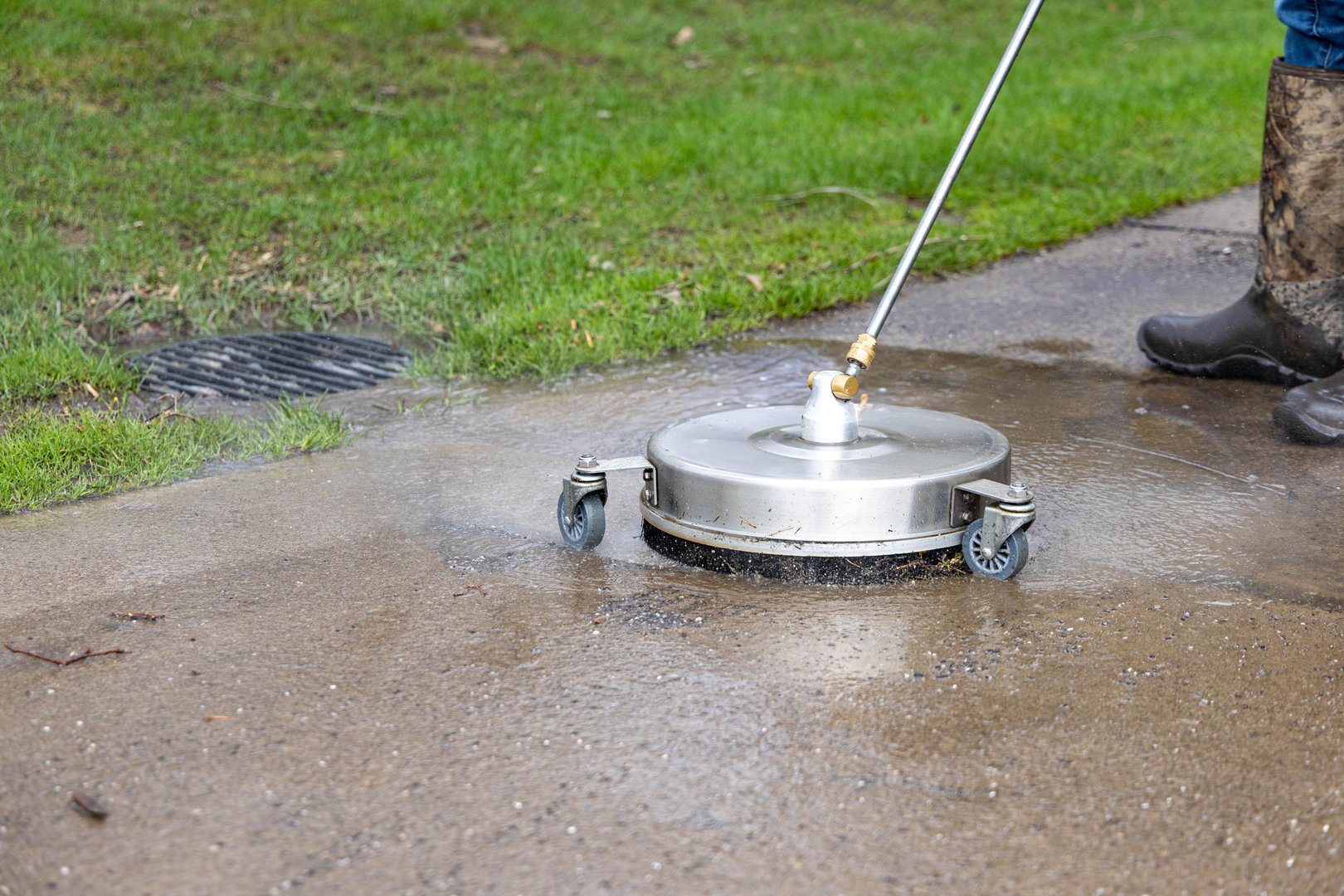 A worker using a surface cleaner to pressure wash a concrete sidewalk with grass on the side.