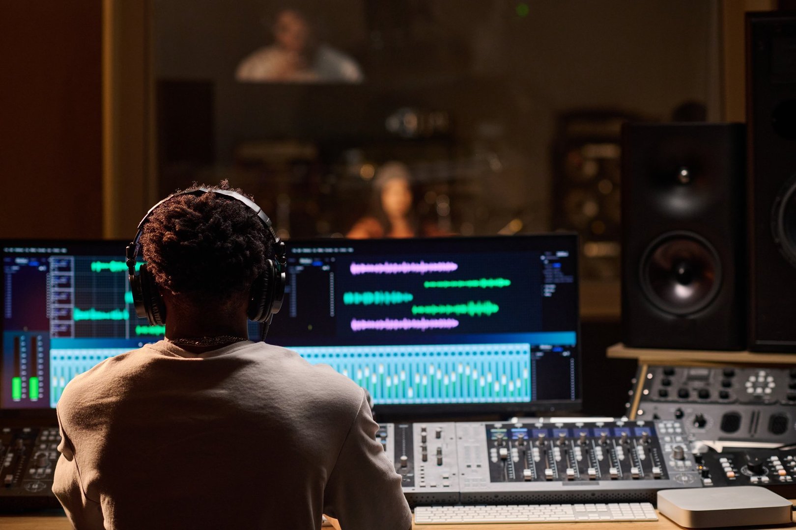 Black man wearing headphones operating audio mixing console and computer monitors in professional recording studio, observing musician performing through glass window in background