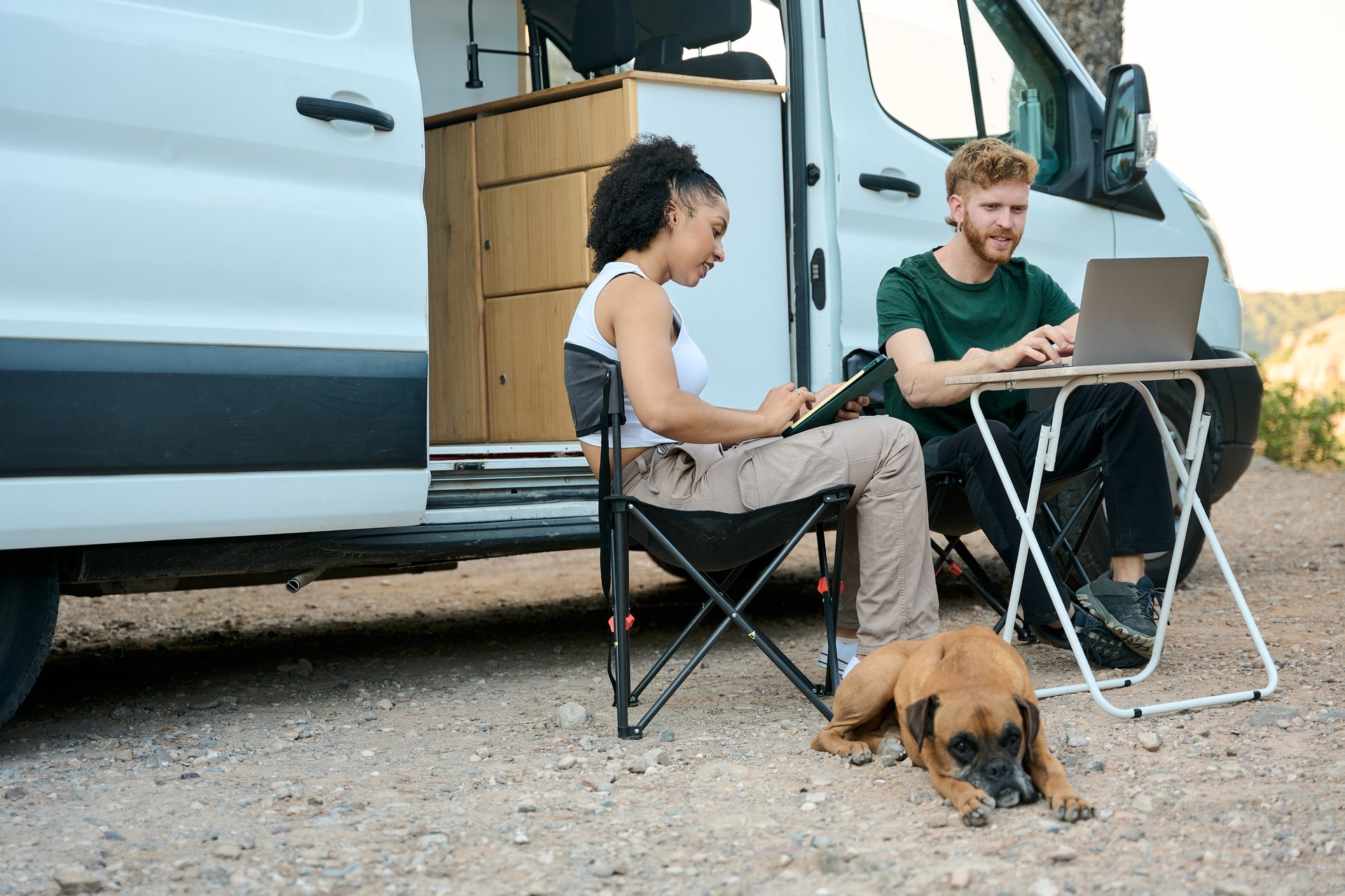 Young multi-ethnic couple working remotely using laptop and tablet near their camper van with their dog