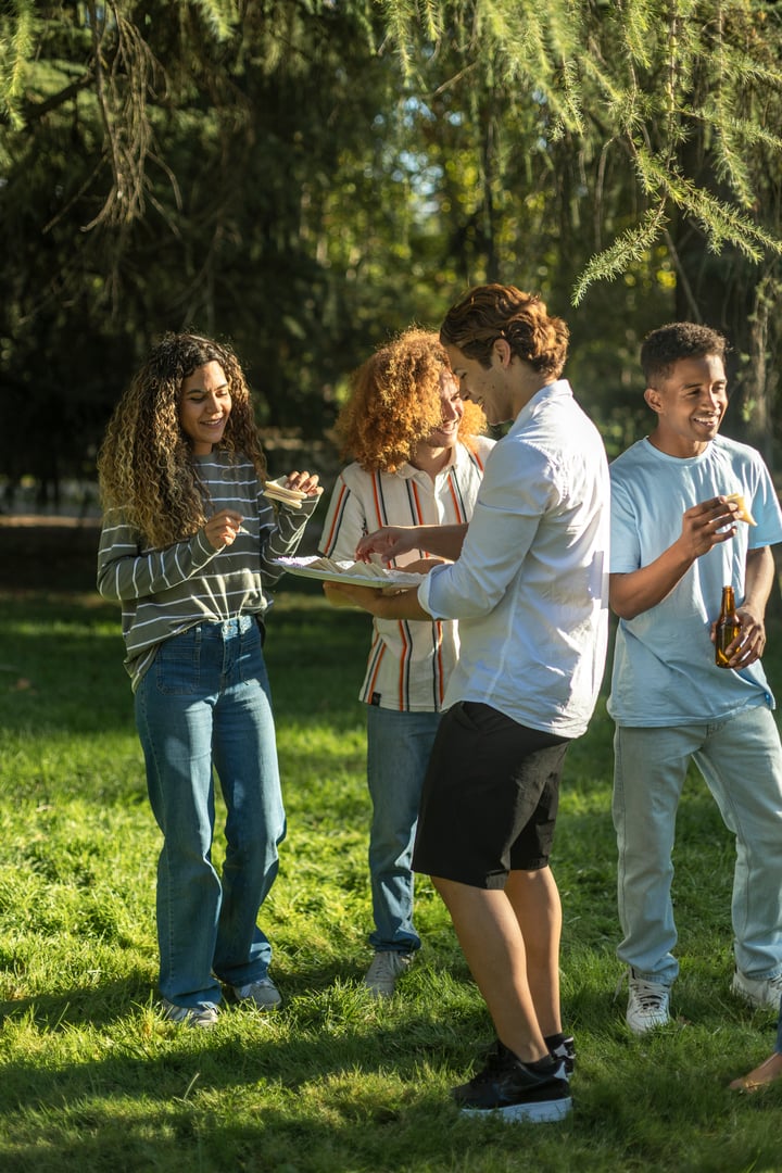 Friends share a lighthearted moment in a park, eating snacks and chatting in the warm sunlight during a casual outdoor event.