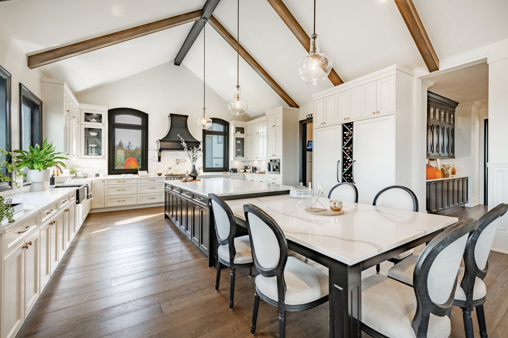 This is a huge kitchen with a modern farmhouse design style the countertops are white granite or marble and the kitchen island is very long and expansive with dining chairs there are houseplants cookbooks small appliances and staging decor on the counters. There is a large wine rack and stainless steel appliances. Hardwood floors wooden beams on the vaulted ceiling, the cabinets are a very dark wood and the windows are trimmed in black with a view to a bright blue sky autumn day showcasing a very colourful scene.