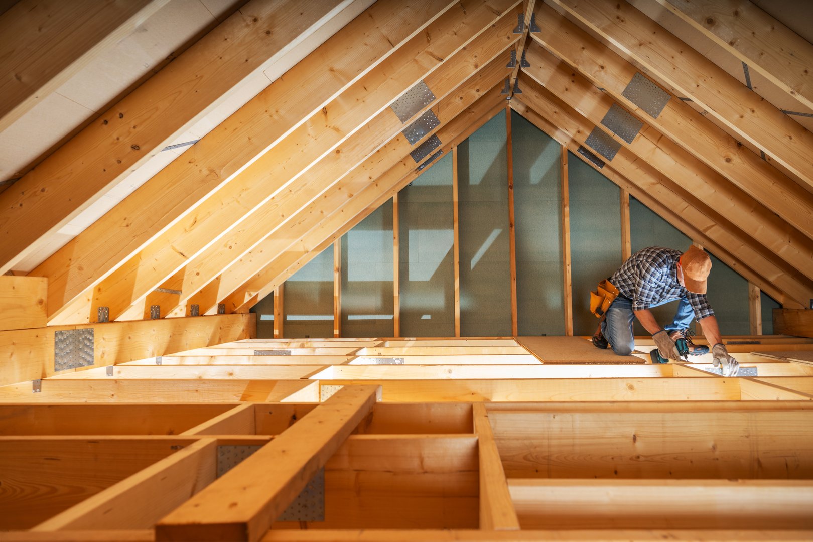A carpenter carefully measures and adjusts wooden beams in an attic, illuminated by sunlight filtering through the roof. The workspace reflects a renovation effort.