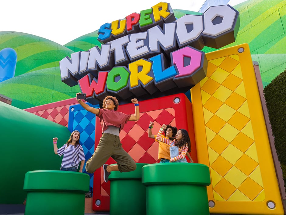 Visitors jump joyfully in front of the Super Nintendo World entrance, featuring colorful pipes and a sign.