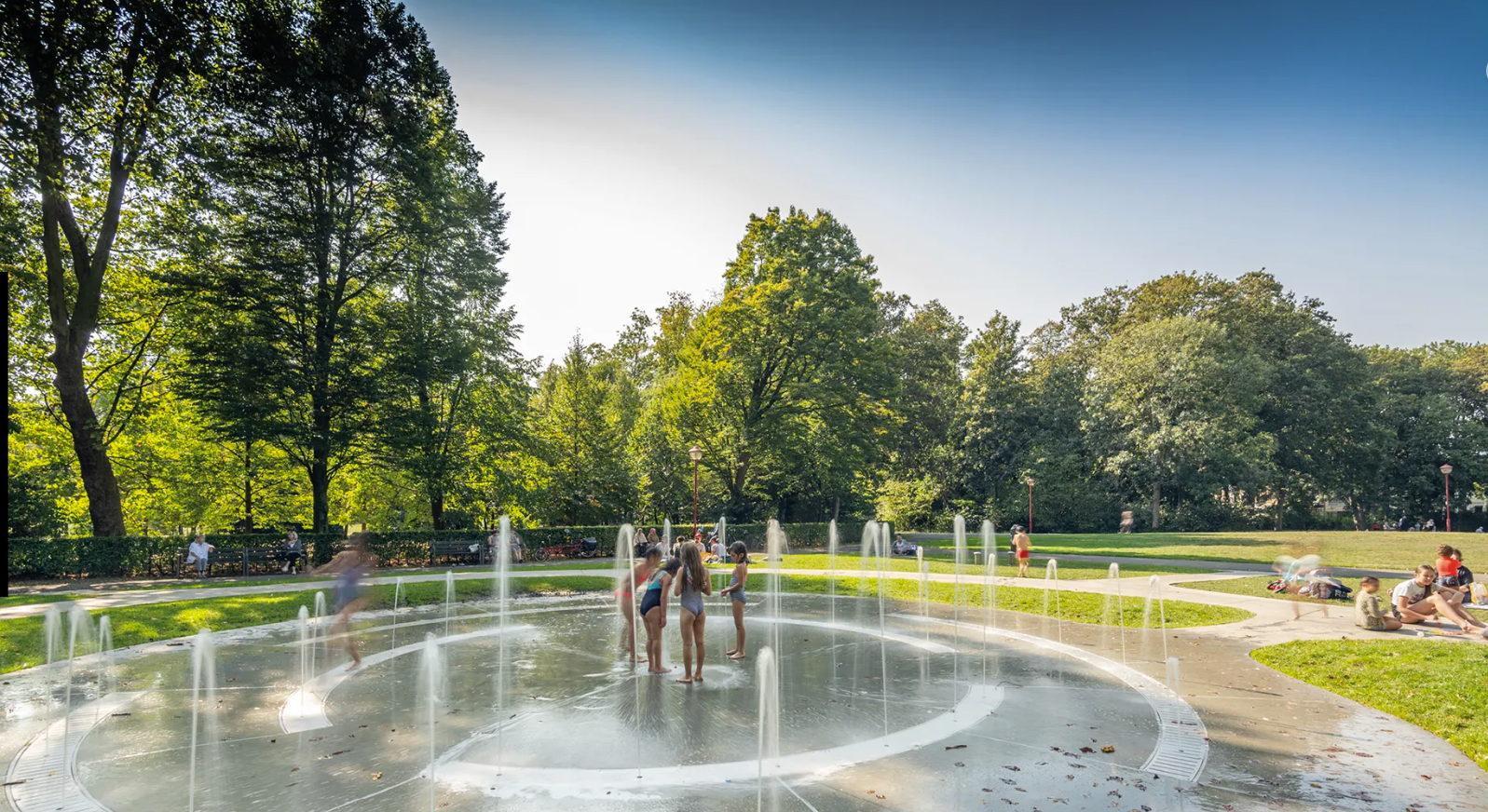 Children play in a circular water fountain in a park surrounded by trees, with people sitting on the grass nearby.