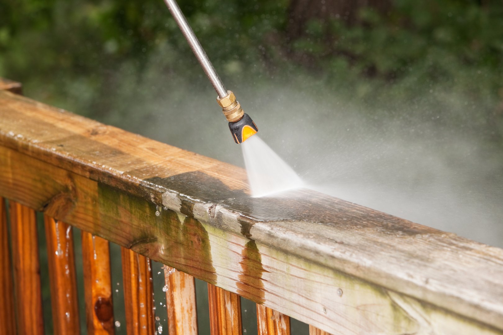 A pressure washer sprayer is cleaning a weathered treated wood deck railing. The wood on the left has been  cleaned, the right is dirty and weathered, showing the contrast. The background are blurred trees.
