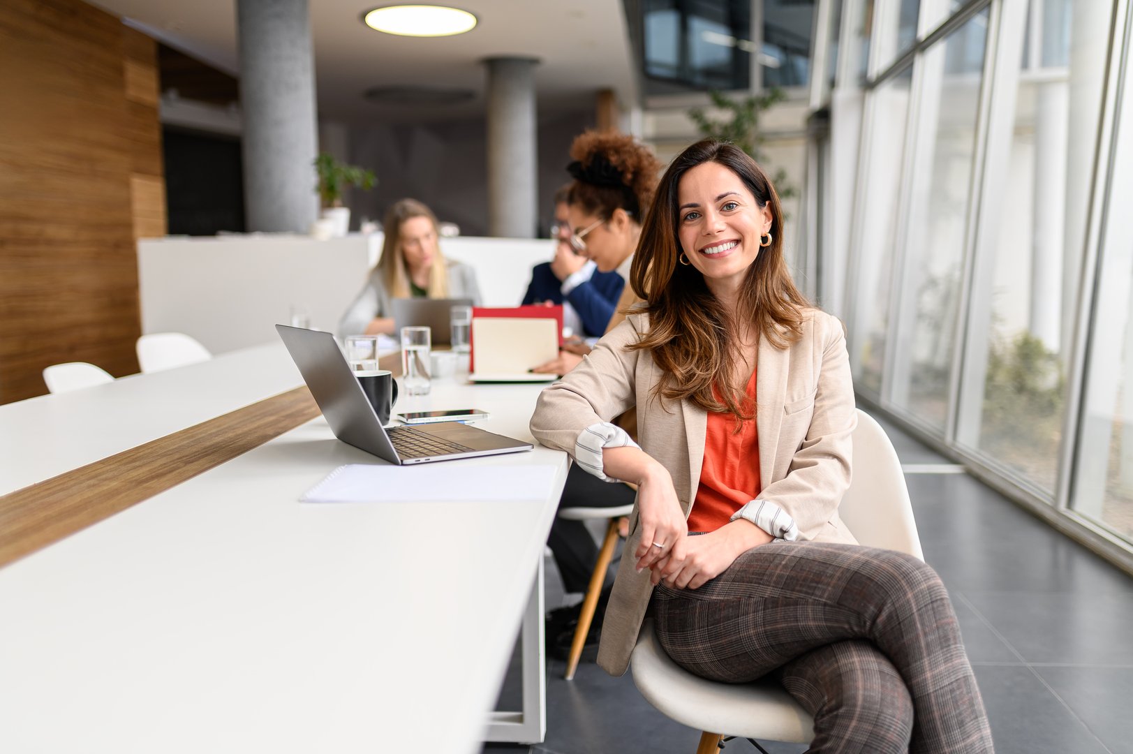 Portrait of female manager with laptop on table working in meeting room while colleagues collaborating in the background