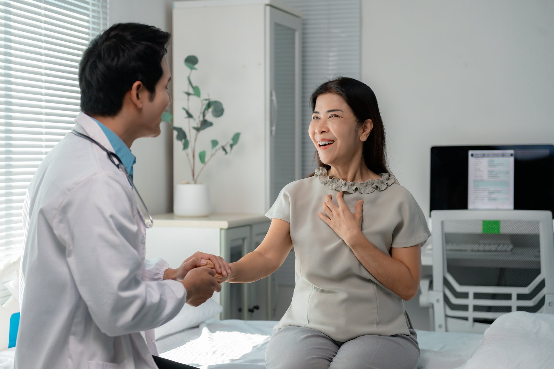 Young male doctor holding hand of a cheerful senior female patient sitting on a hospital bed, providing empathy and support after receiving good news about her health