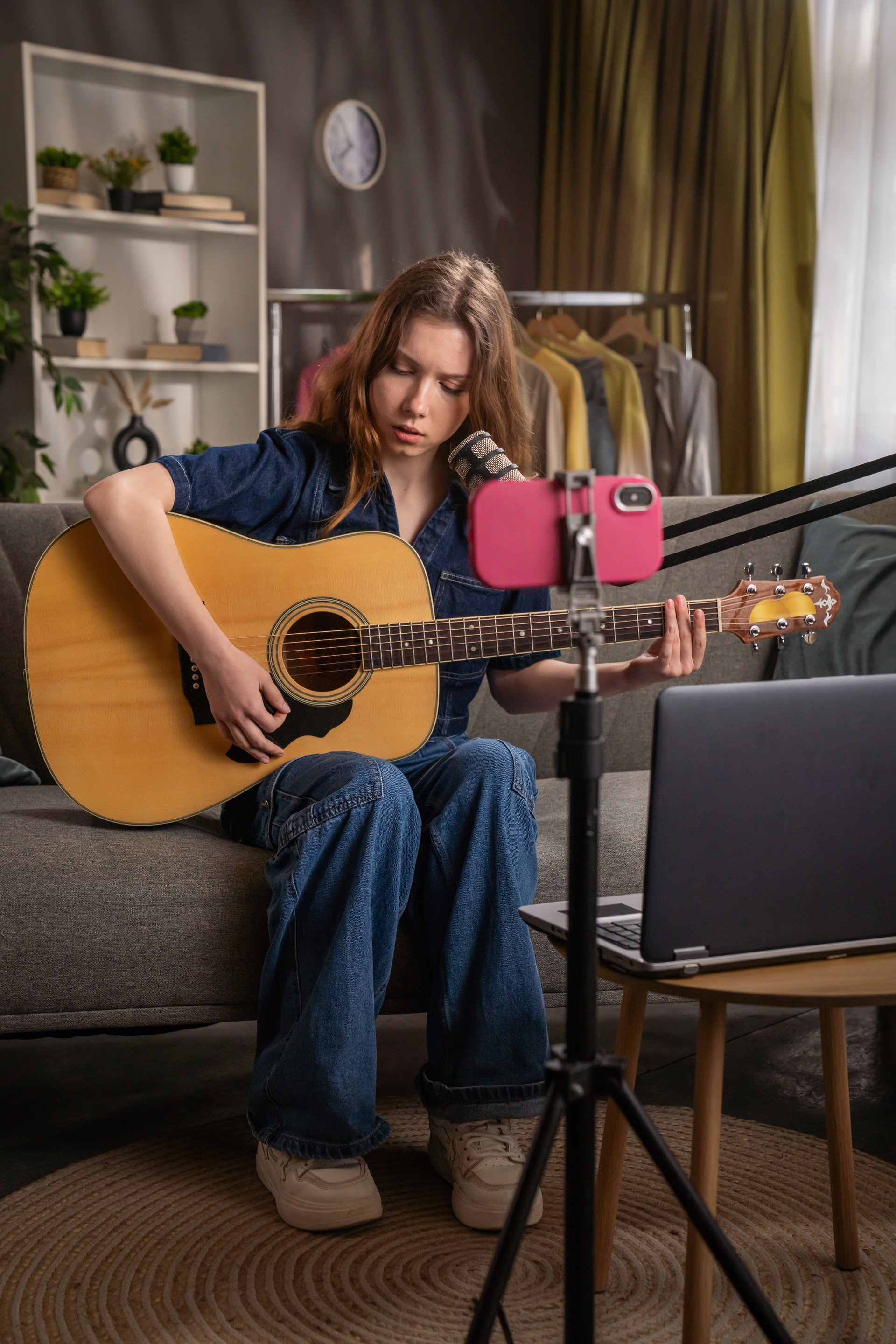 A young woman sits on a couch, playing an acoustic guitar while recording a live music session. Her smartphone and laptop are set up to capture the performance.