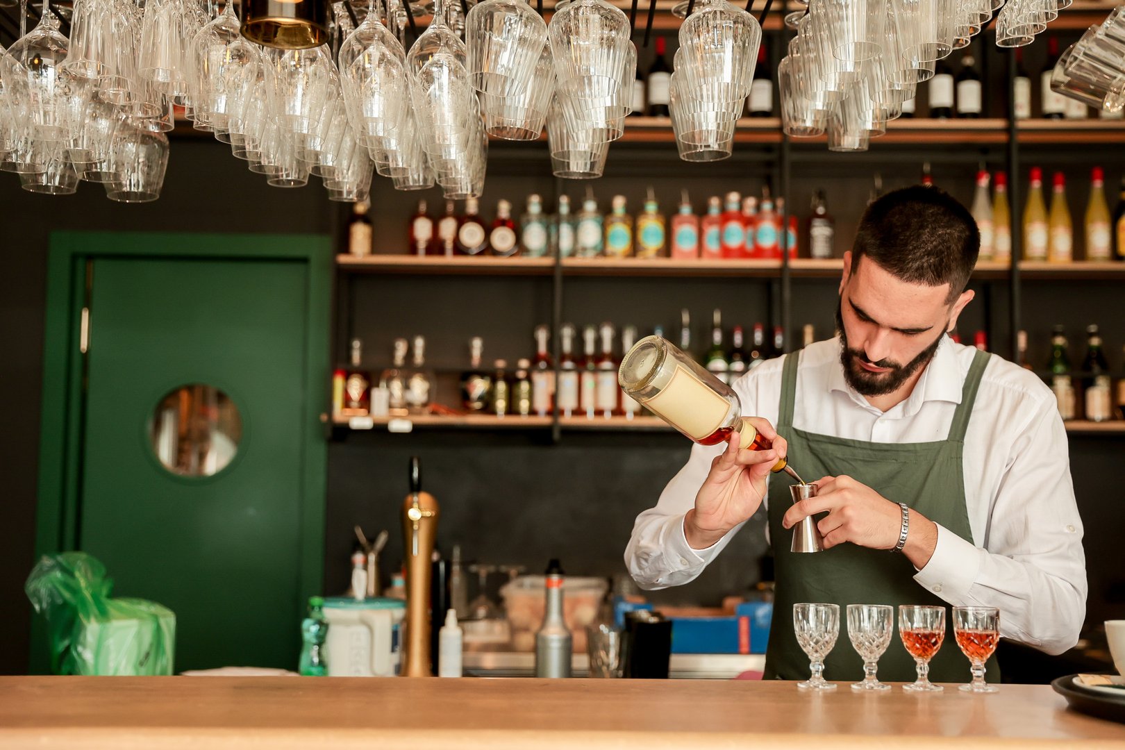 Bartender skillfully pours vibrant cocktails into elegant glasses, showcasing his expertise against a backdrop of shelves stocked with various spirits in a bustling bar