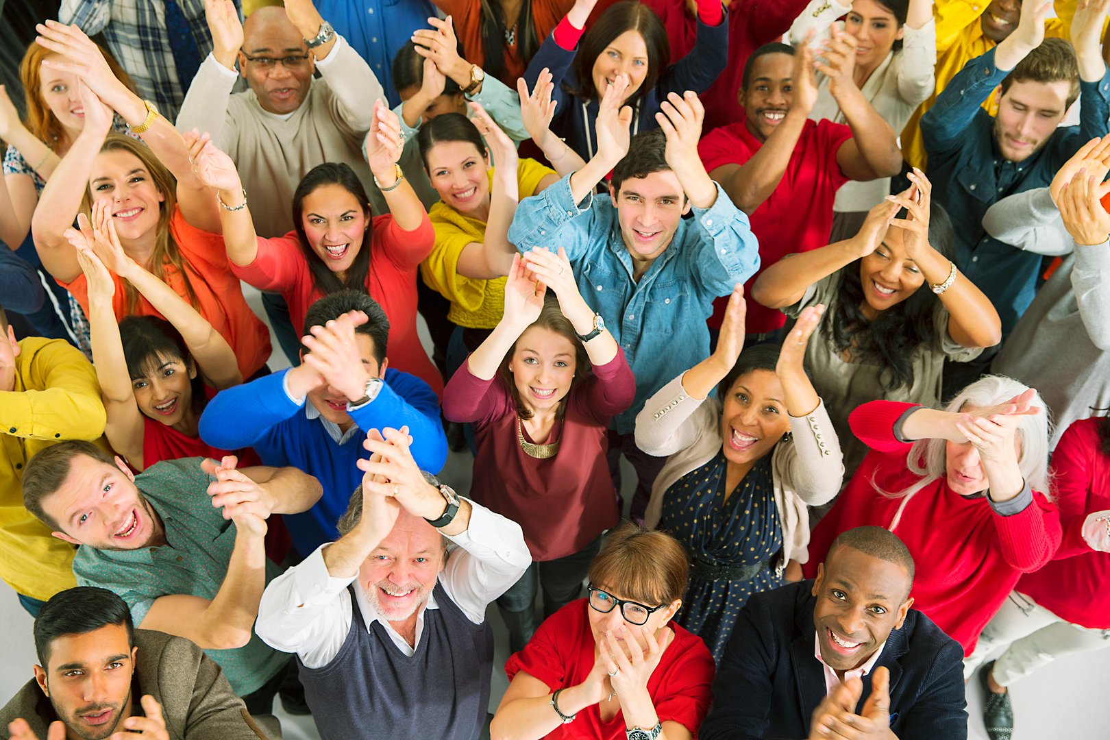 A diverse group of people standing together, smiling, and clapping while looking upward. They appear joyful and enthusiastic.