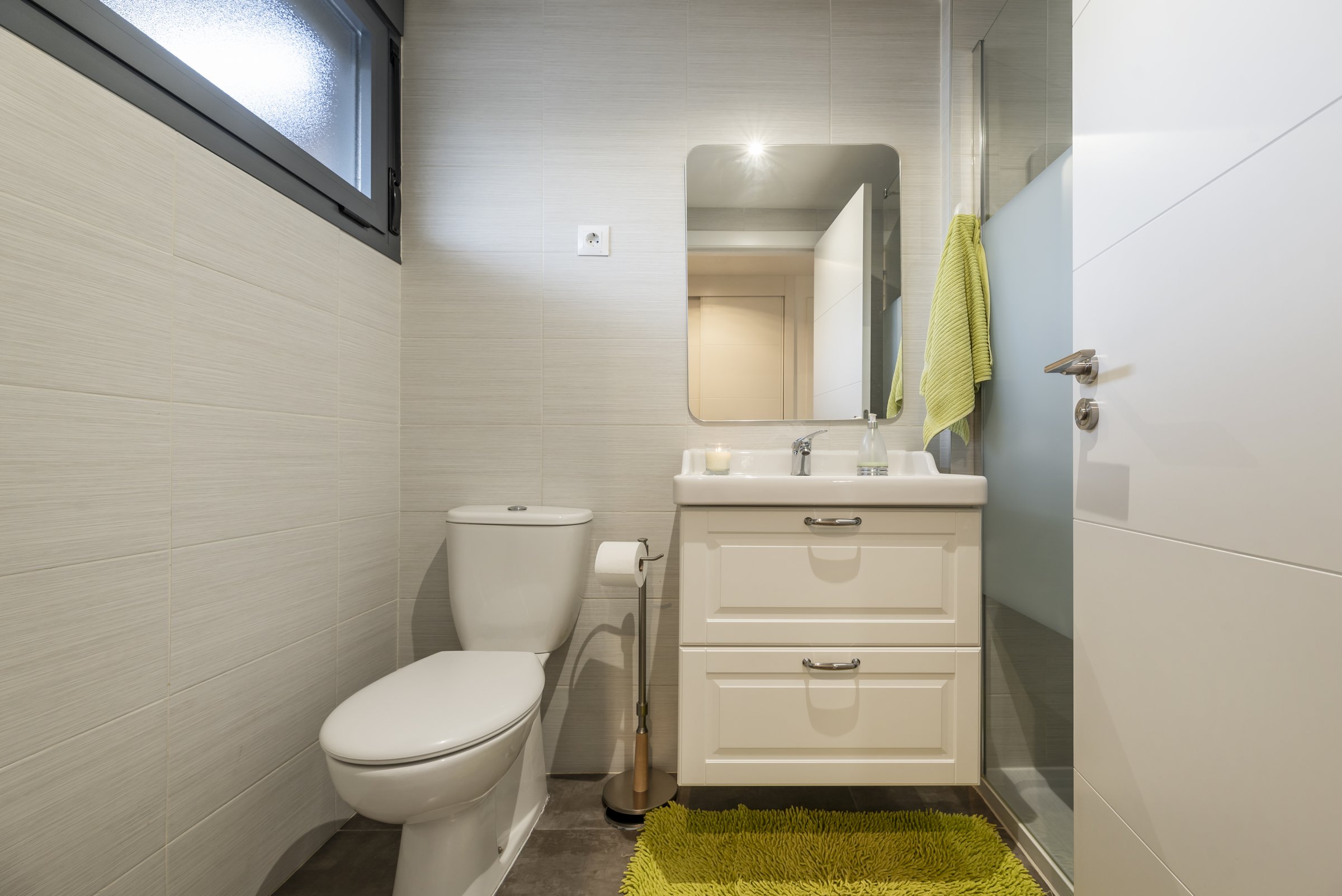 A conventional bathroom with white wooden cabinet, one-piece porcelain sink above the chest of drawers, frameless mirror, green mat and tempered glass screen
