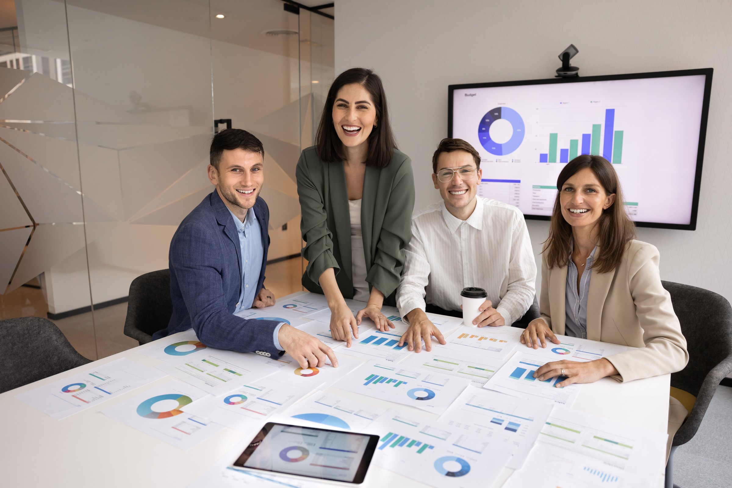 Team of cheerful millennial coworkers of marketing agency staff sitting and standing at large meeting table with paper diagrams, sales reports, posing for company profile portrait in boardroom