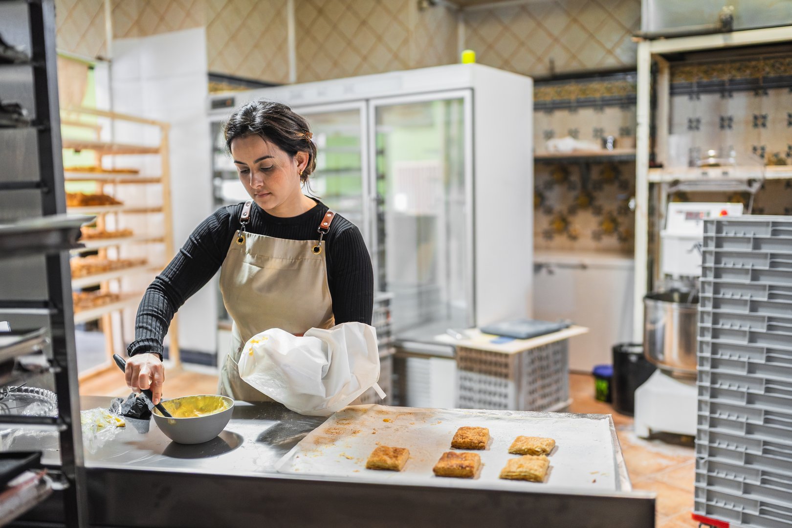 Young woman working as a pastry chef, mixing ingredients in a bowl, preparing to fill pastries in a bakery kitchen