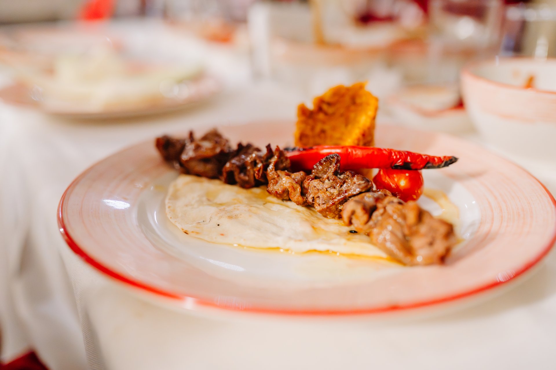 Grilled meat skewer served with flatbread, grilled red pepper, and a side of bulgur or mashed vegetable on a restaurant plate.