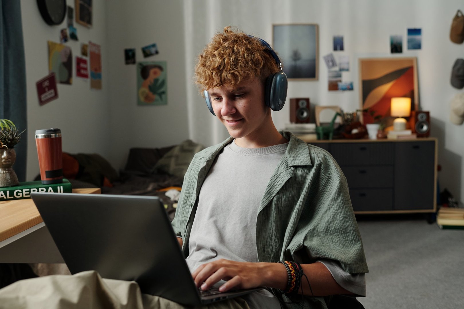 Caucasian teenage boy sitting on bed wearing headphones using laptop smiling engaging with digital content surrounded by personal belongings illustrating gadget addiction concept