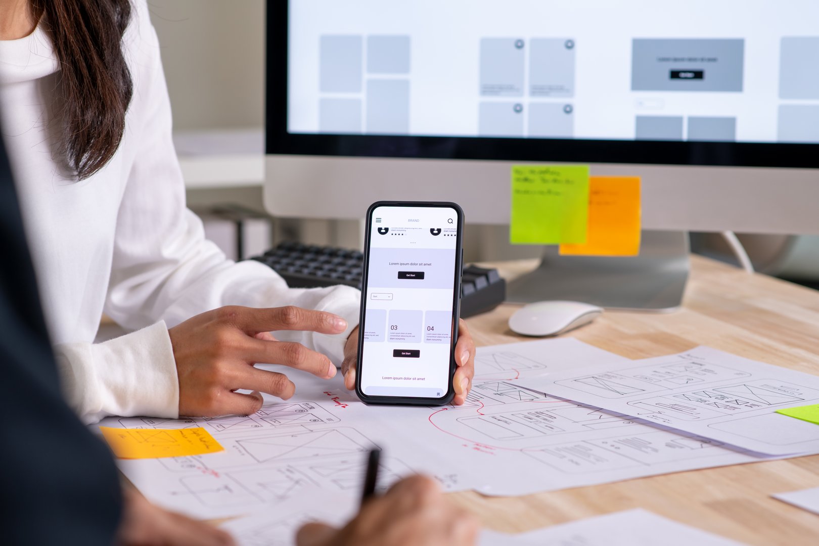 UX UI designer testing prototype on a phone, discussing and brainstorming on wireframes for a website and mobile app prototype, surrounded by sketches of user flow and design tools, in the concept of website and mobile application design concept.
