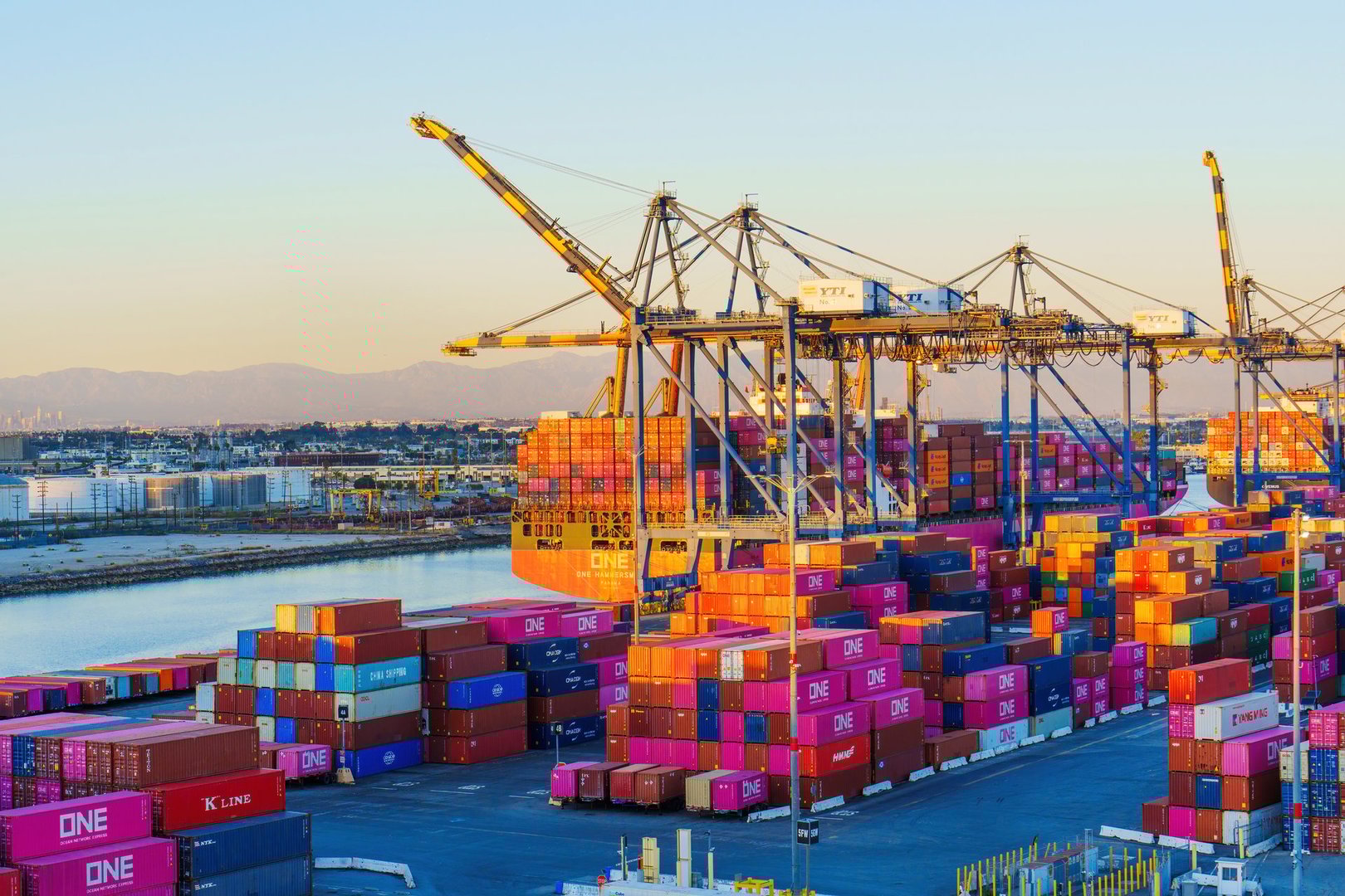 Long Beach, California - January 15, 2025: Dynamic view of cranes loading colorful shipping containers along the waterfront at Long Beach port, with distant mountains visible.