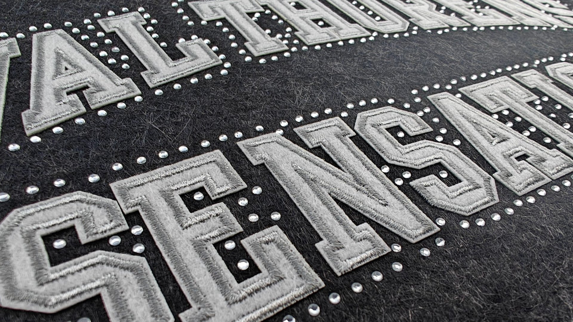 Close-up of embroidered letters SENSATION with decorative stud accents on a dark textured background.