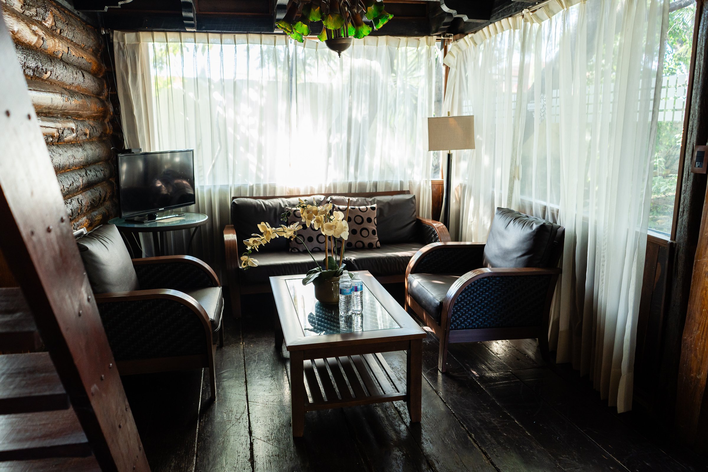 Cozy cabin living room with dark wood, armchairs, sofa, TV, and white curtains.