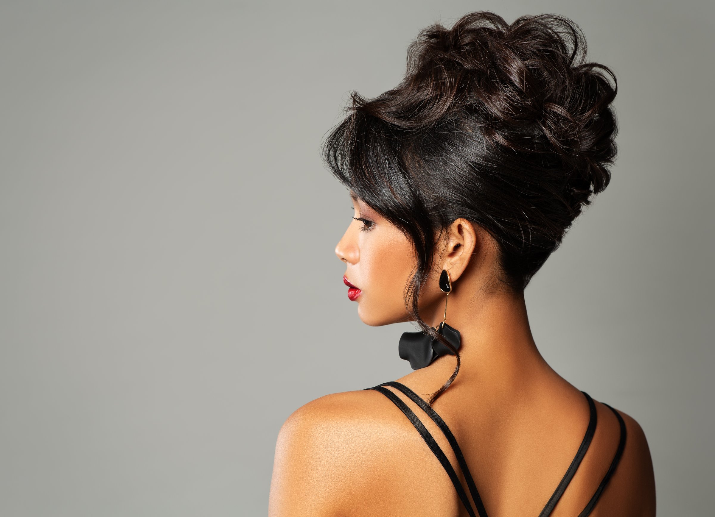 Beautiful Woman with Wedding Updo Hair. Elegant Lady with Evening Hairdo over Gray Background. Fashion Asian Woman with Black Earrings and Red Lips Rear Side View
