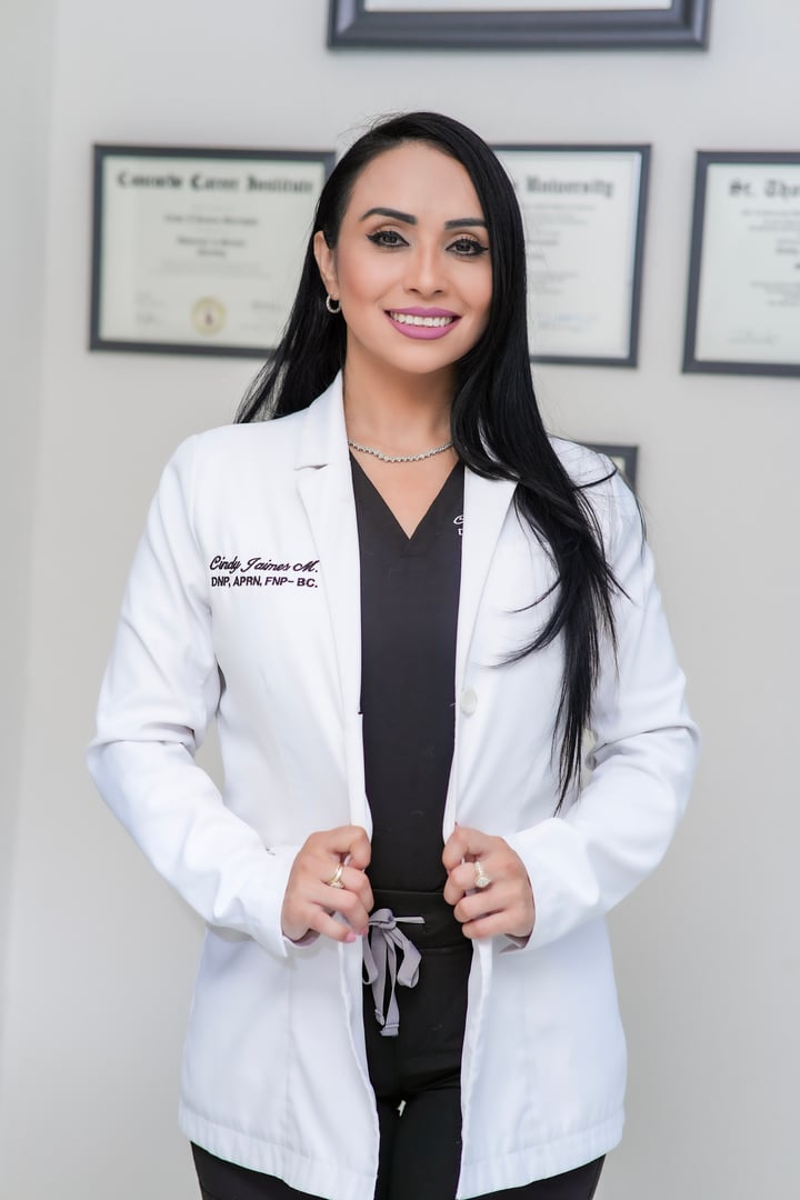 A woman in a white lab coat standing in front of diplomas on a wall, smiling at the camera.