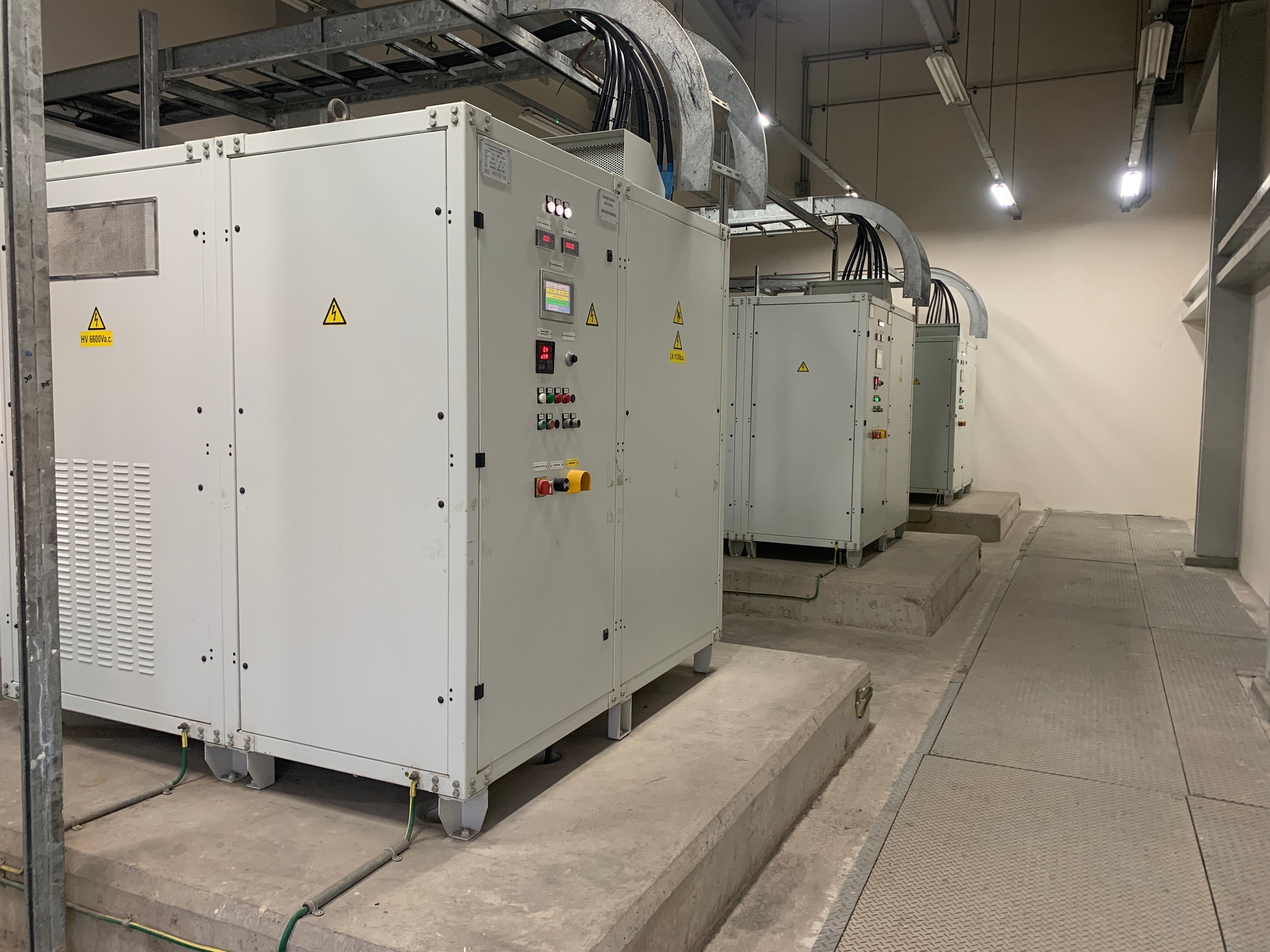 A row of industrial equipment, likely electrical transformers, are lined up in a factory setting. The equipment is white and has control panels with wires and cables running along the ceiling.