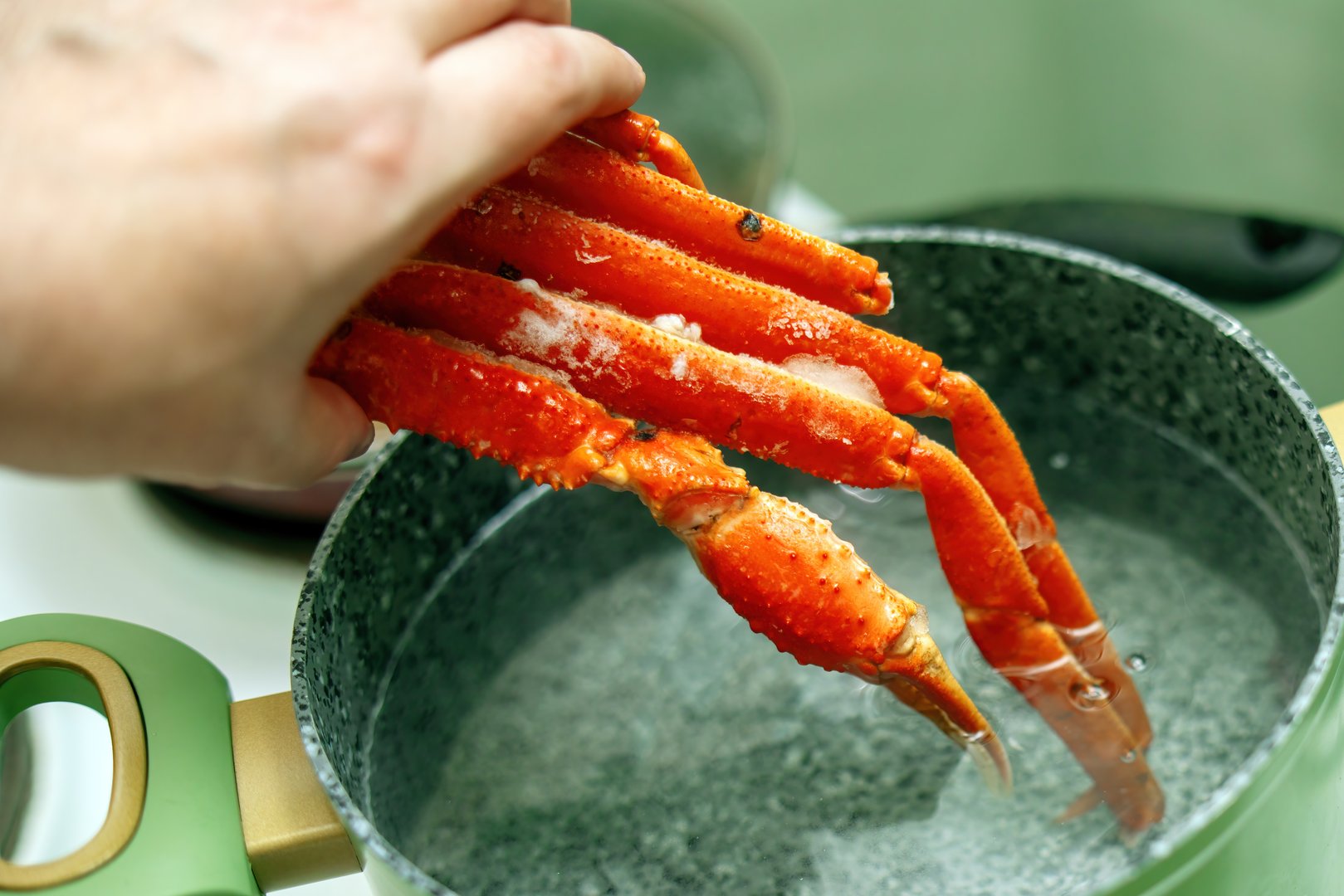 person's hand places frozen cooked crab legs into a green pot filled with water for reheating. The scene showcases a culinary preparation process