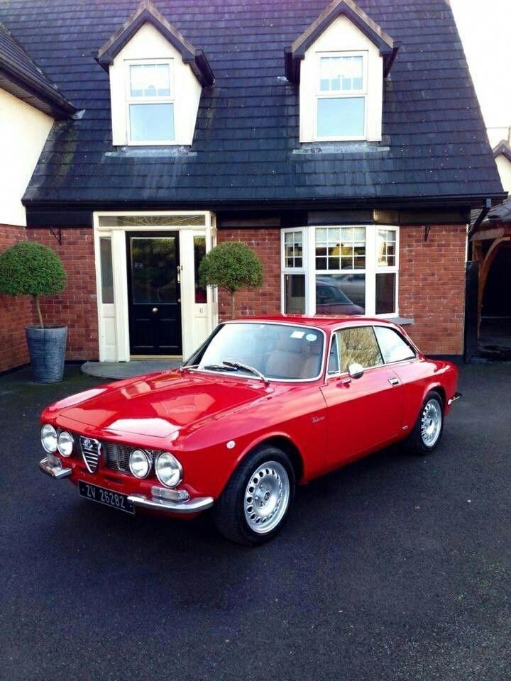 Red vintage Alfa Romeo car parked in front of a house with dormer windows and topiary trees.