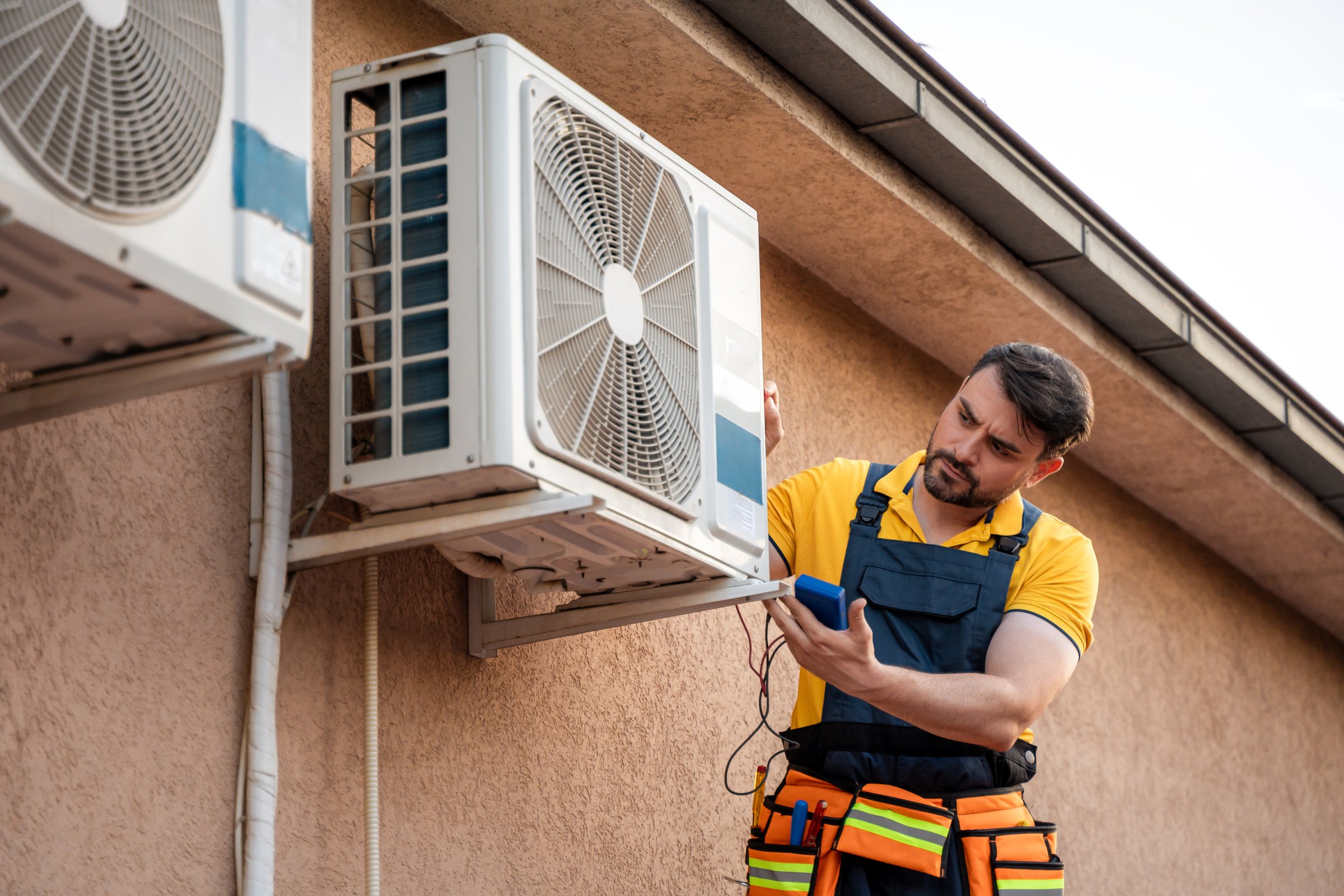 A technician is checking an outdoor air conditioning unit while standing on a ladder at a home. The sun is shining, and the day looks clear.