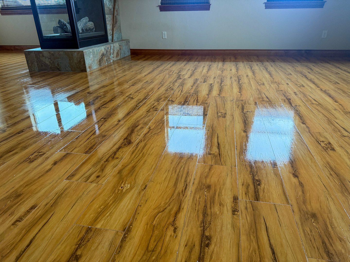 Shiny hardwood floor in an empty room with natural light coming through windows. The polished wooden flooring shows reflective patches where sunlight hits the surface. The room features white walls and a stone or marble fireplace base visible in the corner. The warm-toned wood planks display natural grain patterns and knots throughout the floor surface.