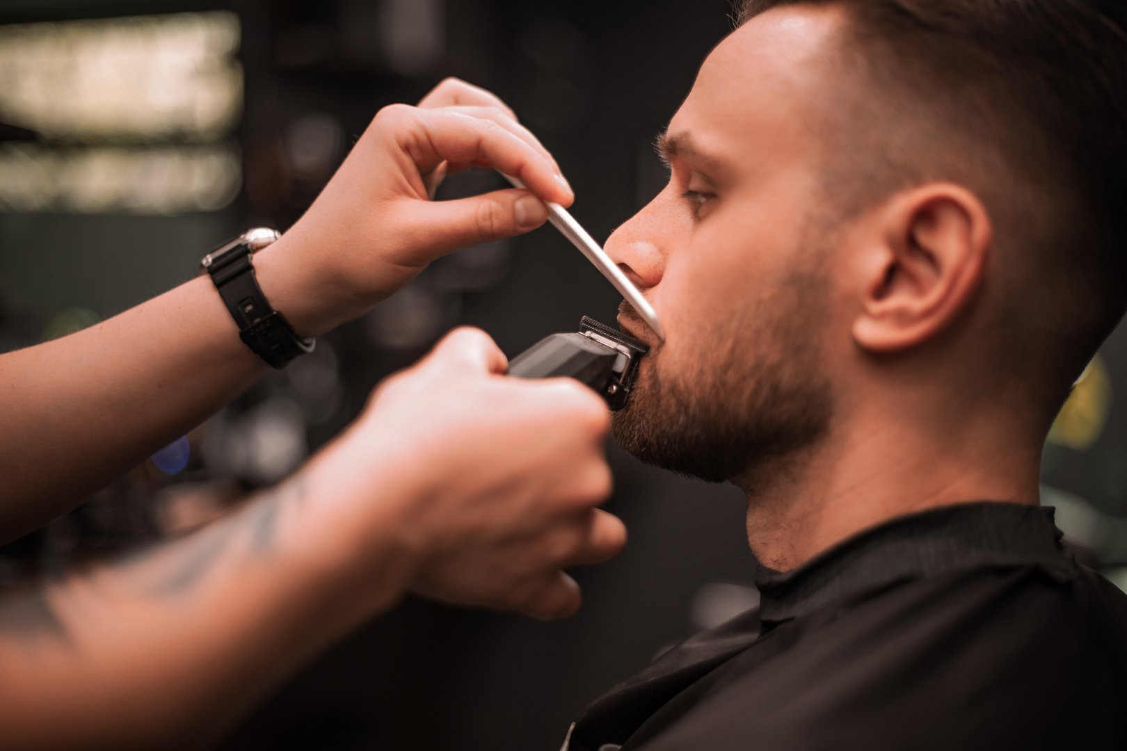 Close-up of a barber trimming a client's mustache with electric clippers and a comb, showcasing precise grooming technique and personal care in a modern barbershop setting.