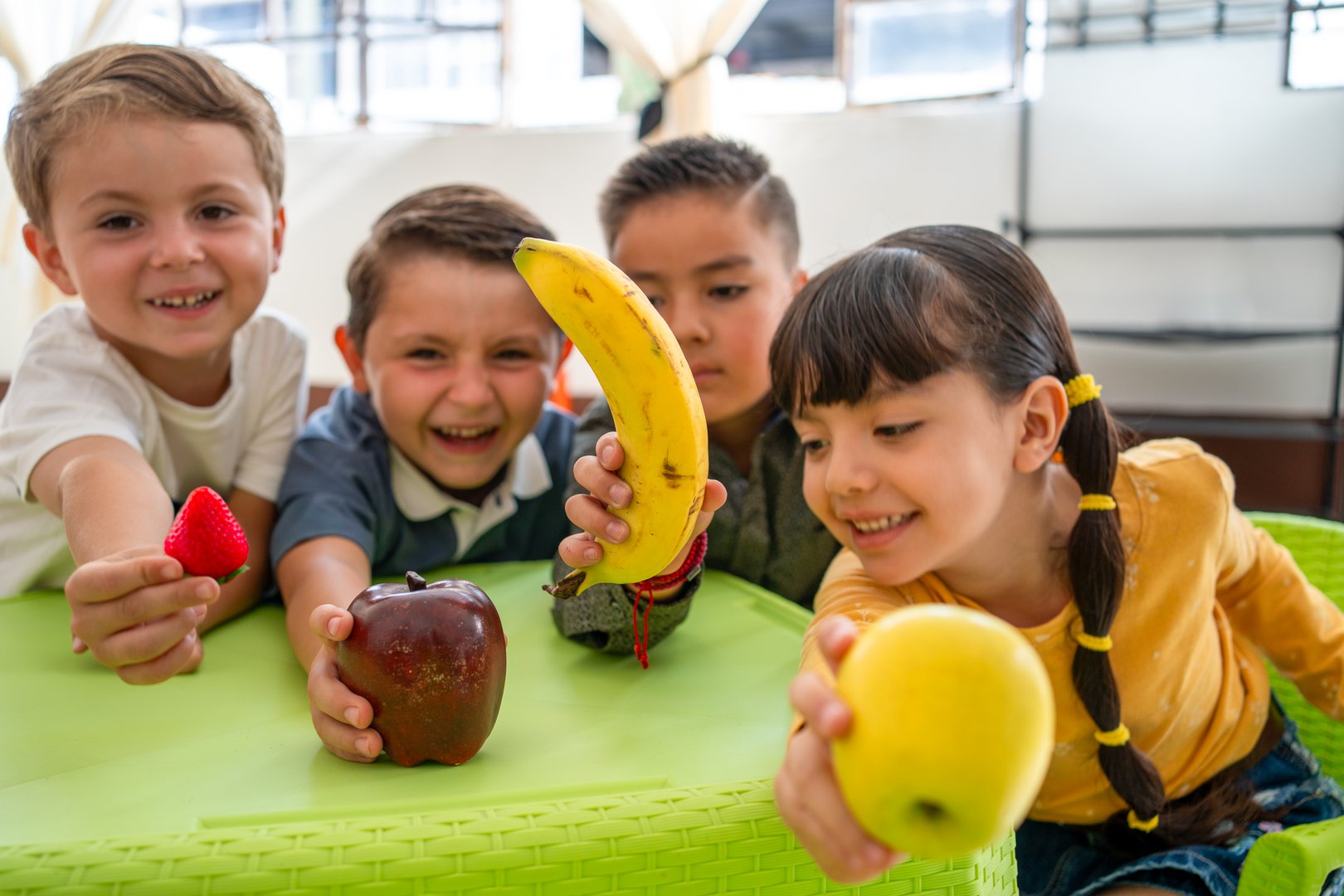 Diverse kindergarten children smile and show fruits inside a classroom. Concept of healthy eating, nutrition habits, and fruit awareness during lunch or educational activities