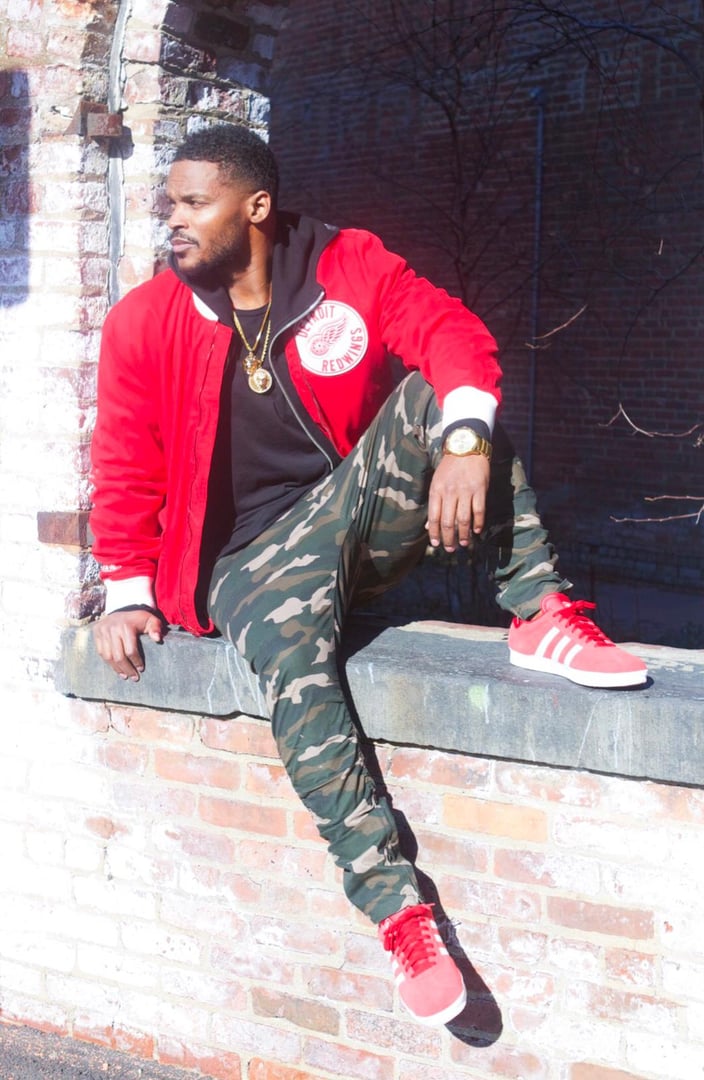 Man in red jacket and camouflage pants sitting on a brick ledge, wearing red sneakers and gold accessories.