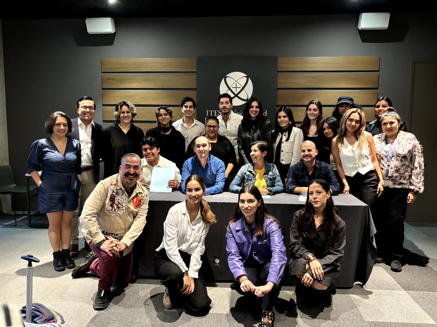 A group of people posing together indoors with a logo in the background, some seated at a table, others standing behind.