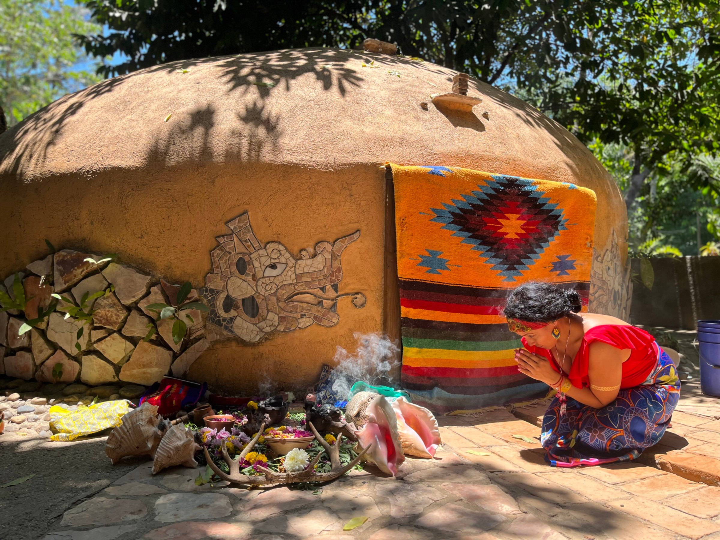 A person in vibrant clothing praying outside a decorated dome structure, surrounded by offerings on a sunny day.