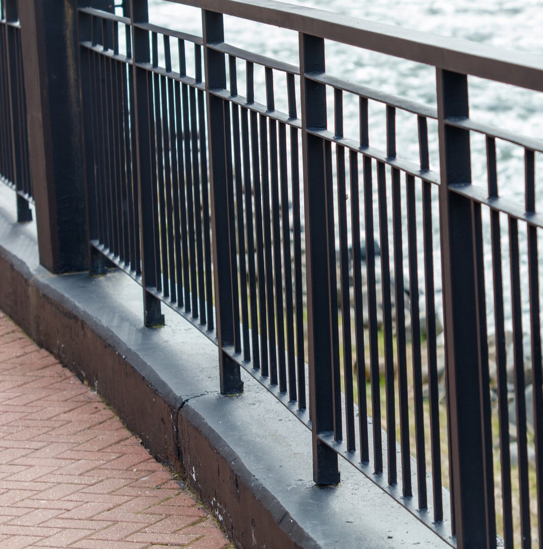 A black metal fence with a brick wall behind it. The fence is on a walkway next to a body of water