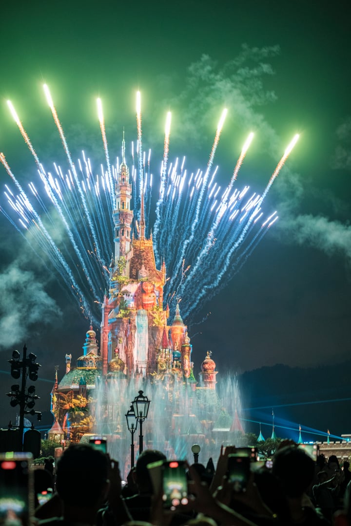 Spectators gather in awe as vibrant fireworks burst into the night sky, reflecting off the beautifully lit castle at Disneyland Hong Kong.