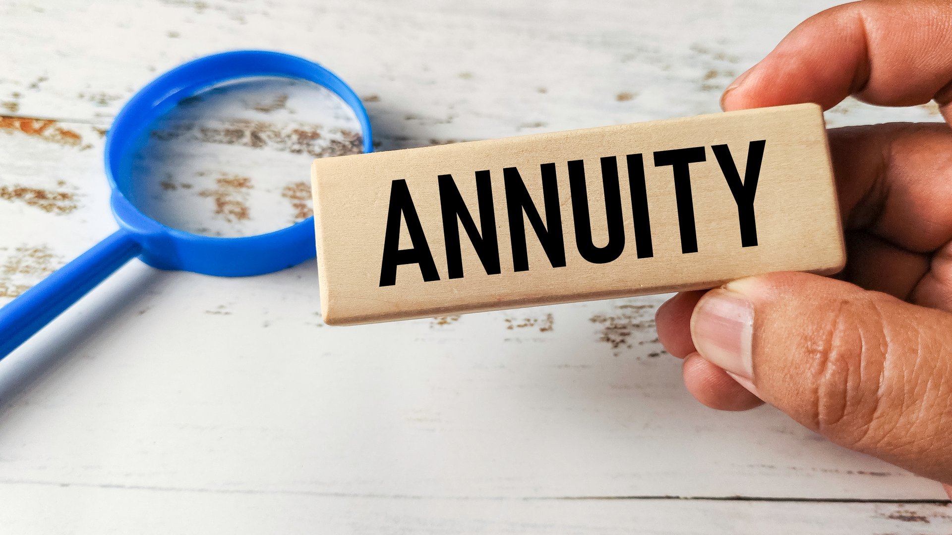Hand Holding a Wooden Block Engraved with Annuity Next to a Magnifying Glass, Representing Retirement Savings and Financial Planning