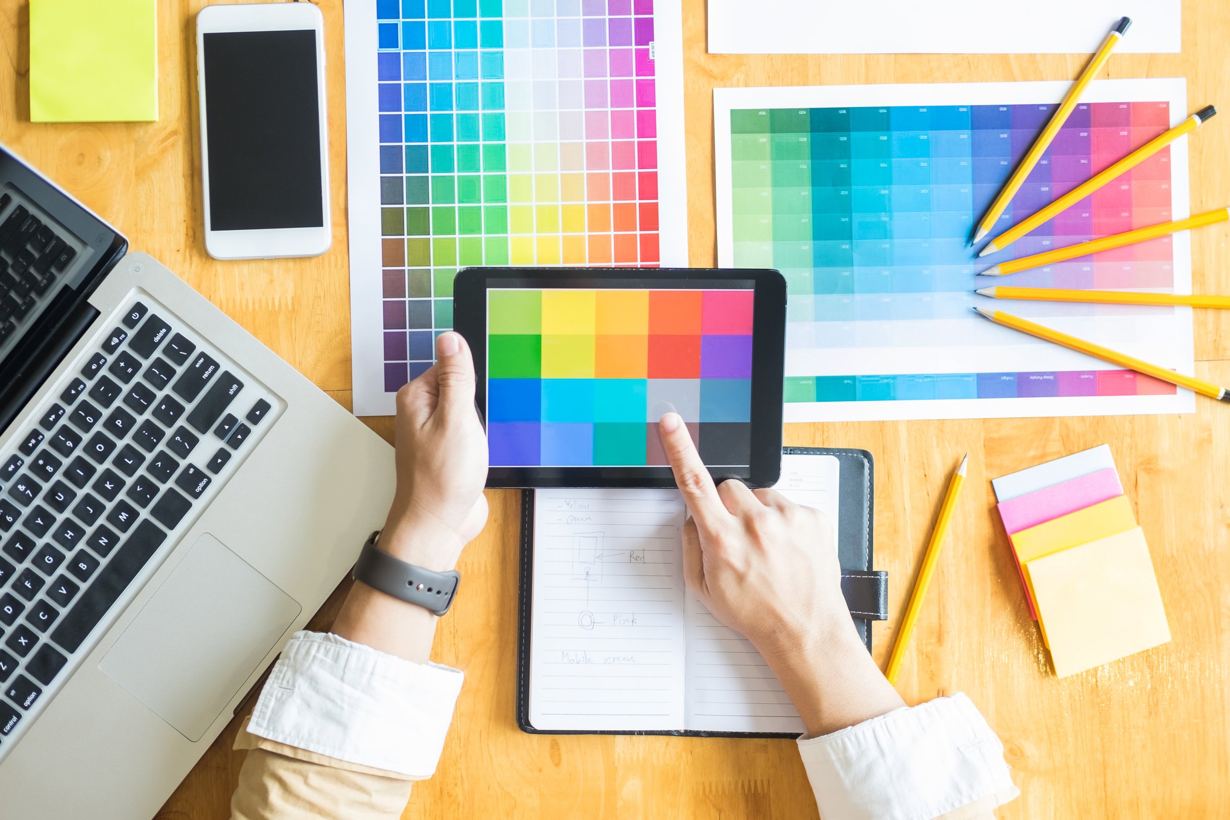 Person selecting colors on a tablet surrounded by color swatches, a laptop, smartphone, notebook, and pencils on a desk.