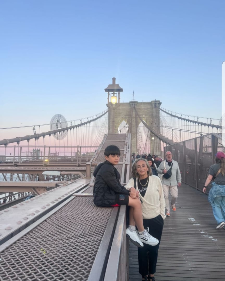 Two people on a pedestrian walkway of the Brooklyn Bridge at sunset. The bridges towers and cables are visible in the background.