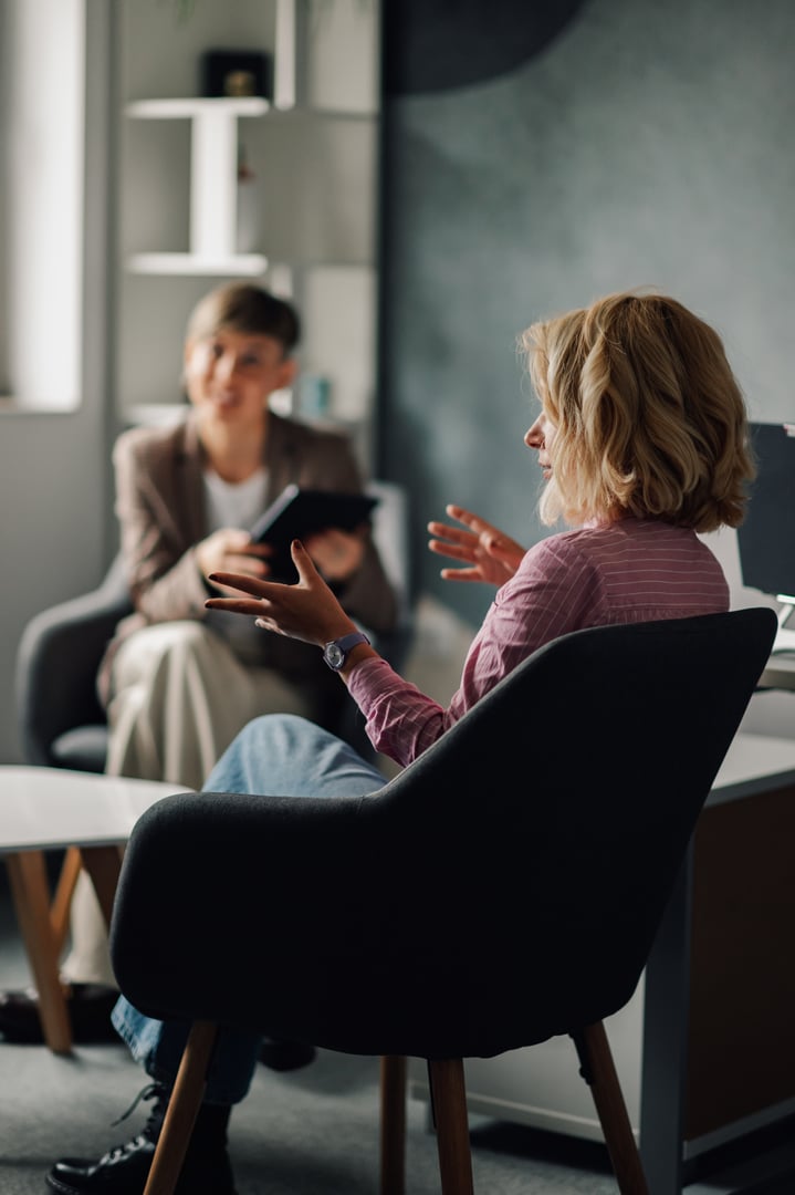 Woman sitting comfortably in an armchair, engaging in conversation with a psychologist who is taking notes on a digital tablet during a therapy session in a modern office setting