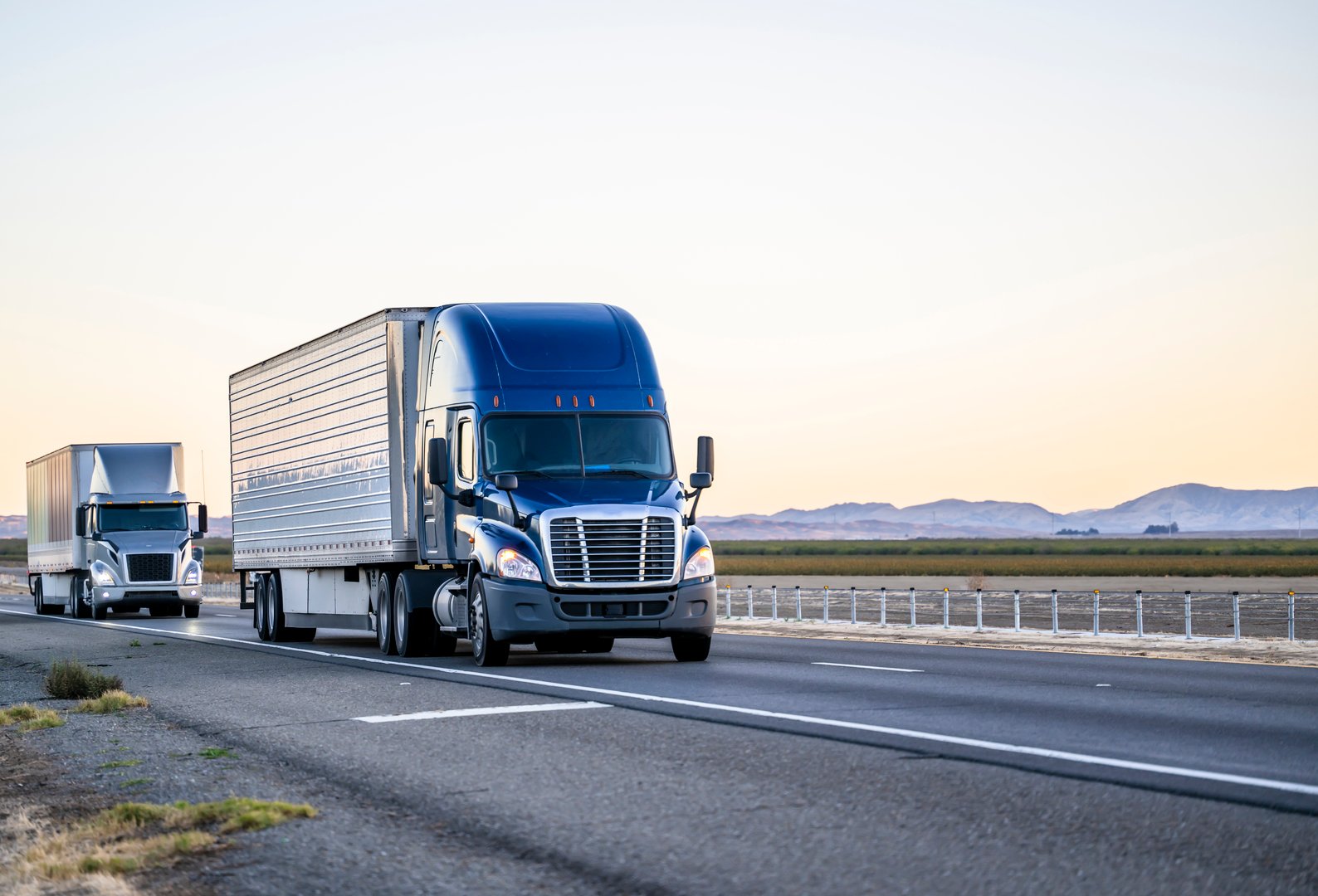 Industrial commercial big rigs long hauler semi trucks with semi trailers transporting different cargo driving on the wide divided straight highway road between the fields in California