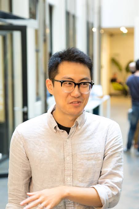 Person wearing glasses and a light shirt, standing indoors, gesturing with one hand, in a brightly lit hallway.