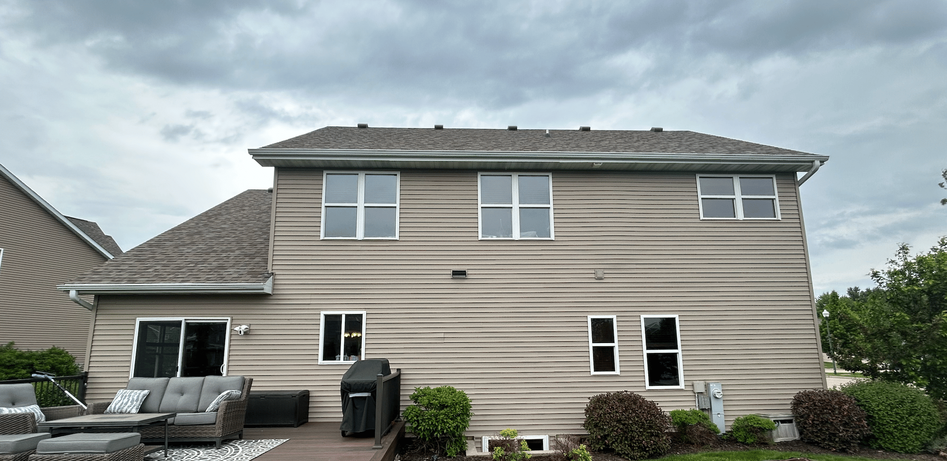 Back view of a two-story house with tan siding, double windows, a deck with patio furniture, and overcast sky.