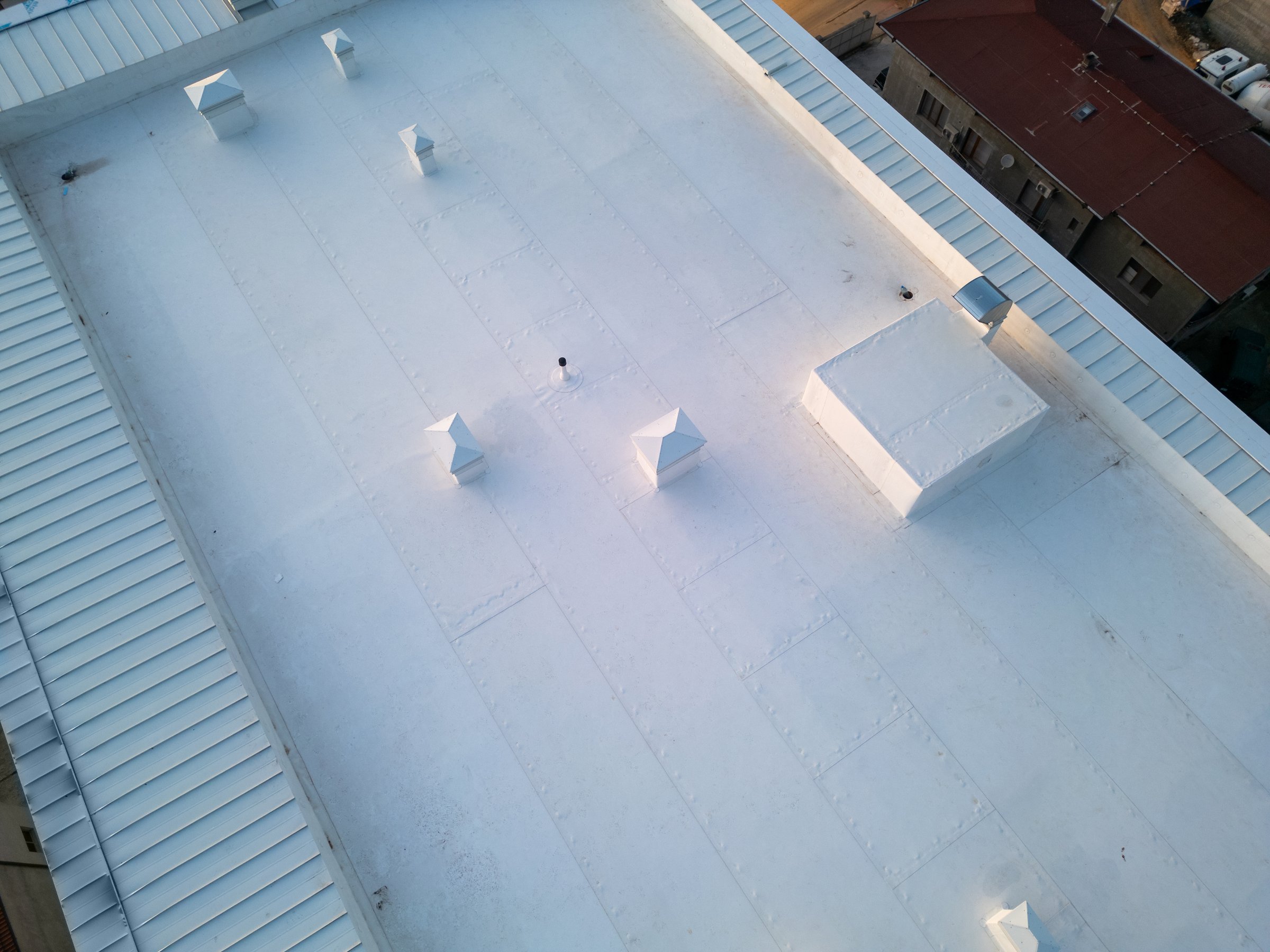 Aerial view of a white industrial rooftop showcasing a clean, minimalistic design with multiple vents and structures. The symmetrical layout and geometric patterns create a modern architectural aesthetic, captured from a drone perspective.