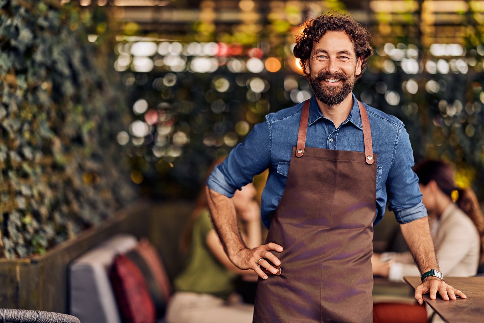 Cafe owner wearing an apron, smiling warmly, standing confidently in outdoor setting.