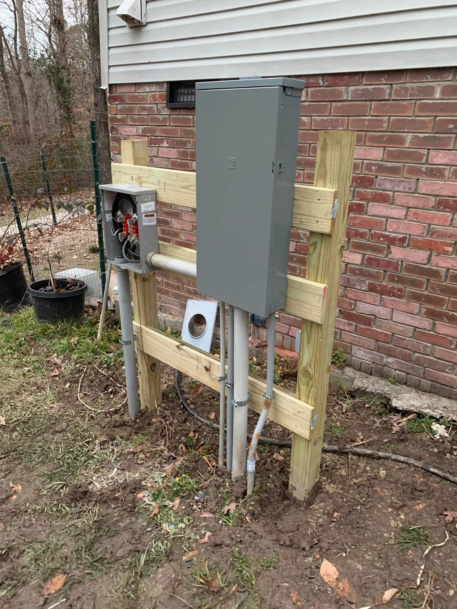 Electrical panel and meter setup mounted on wooden posts near a brick house, with conduit pipes below.