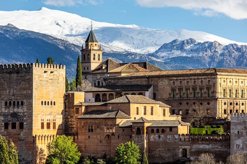 View of the Alhambra in Granada, Spain, with snow-capped mountains in the background under a clear blue sky.