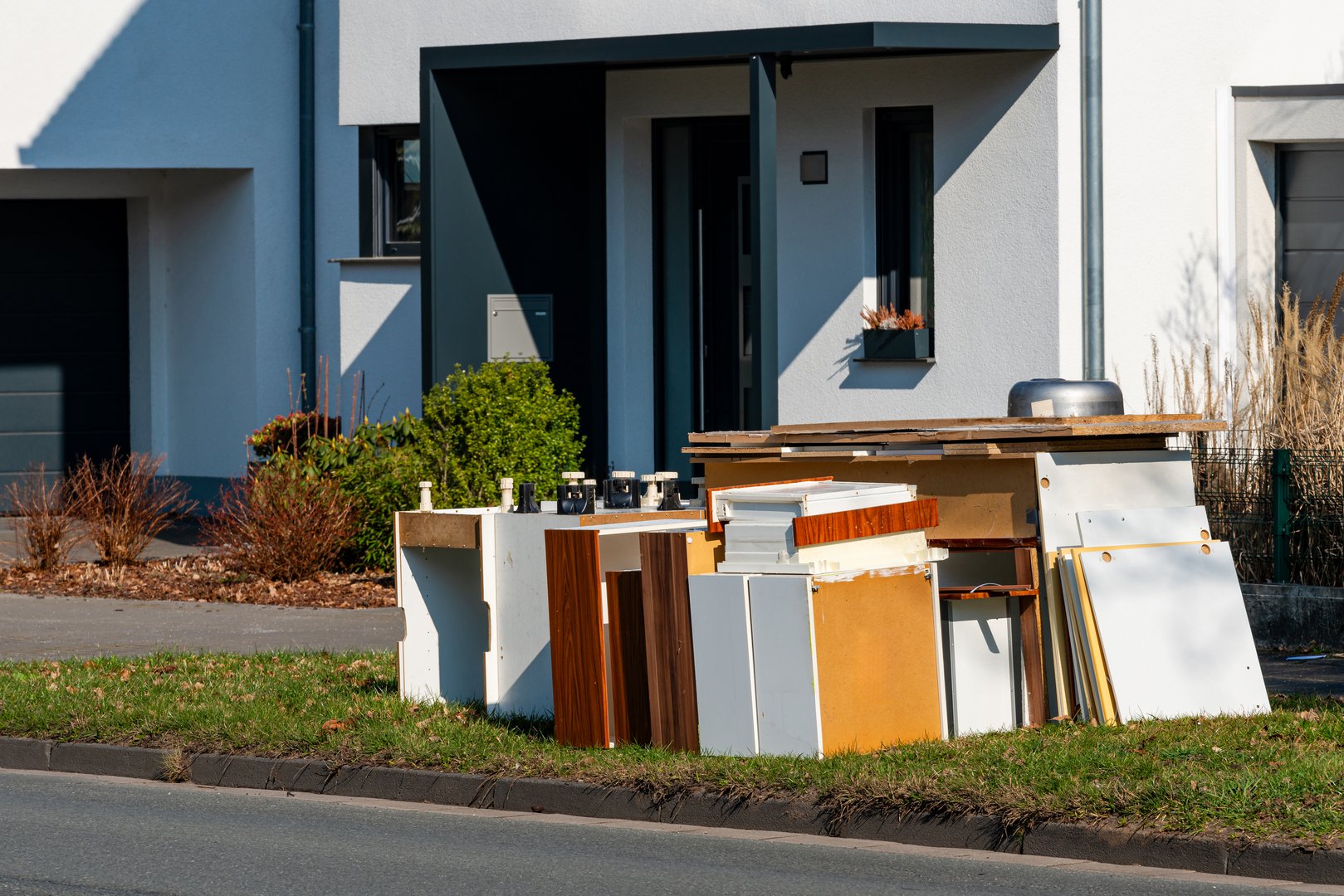 A collection of discarded furniture pieces and wooden materials sits on the grass beside a modern house. The sun shines brightly, highlighting the textures and colors of the items.