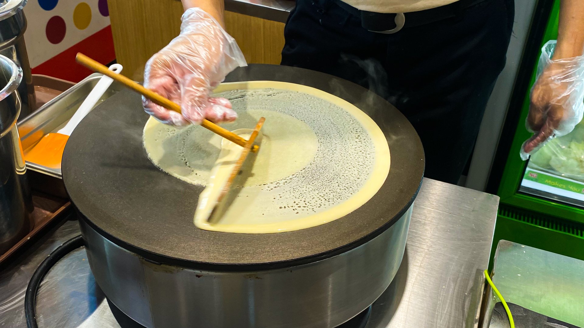 A person is making a crepe on a hot plate using a wooden utensil.